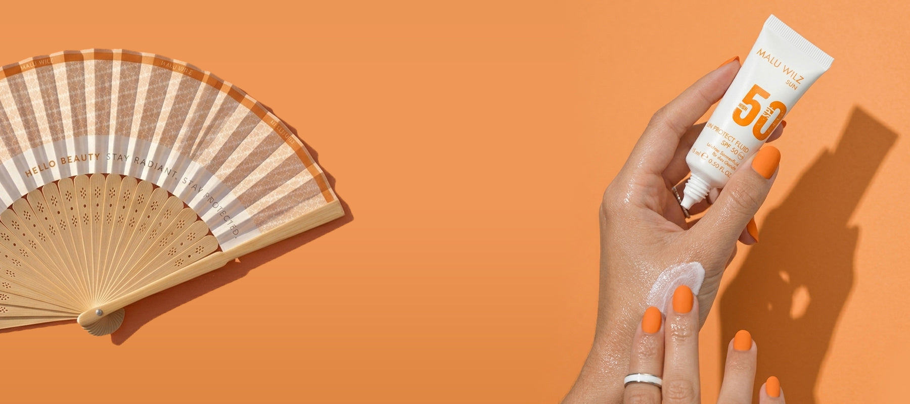 Eine Hand trägt eine Sonnencreme auf den Handrücken auf, daneben liegt ein dekorativer Fächer in Orangetönen vor einem einheitlich orangefarbenen Hintergrund.