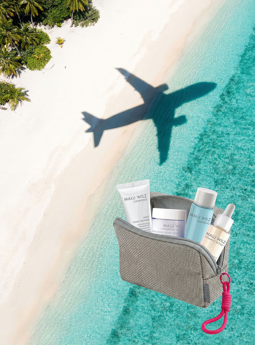MALU WILZ Travel Size Promo: Kosmetiktasche mit Reisegrößen neben einem tropischen Strand mit Flugzeugschatten.