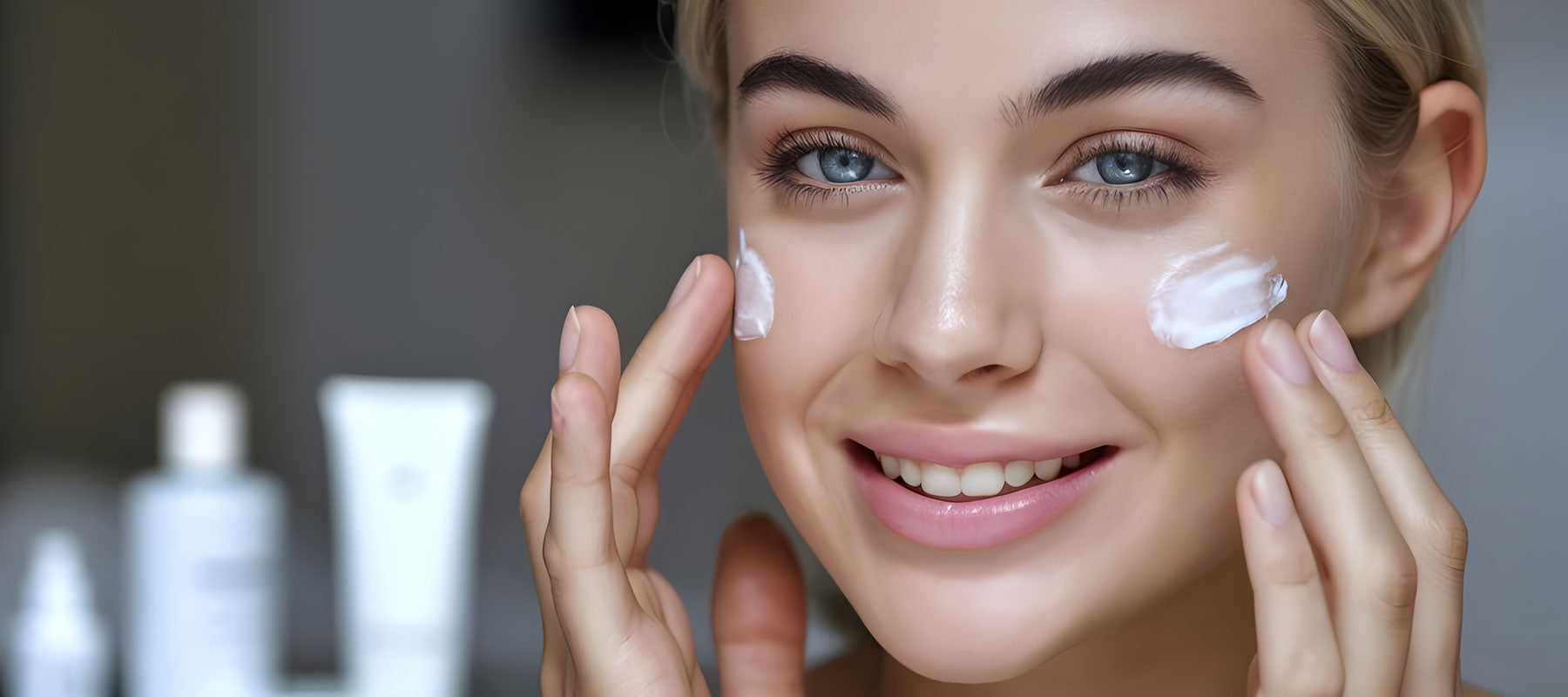 Nahaufnahme einer lächelnden Frau, die sich Creme auf ihre Wangen aufträgt, mit unscharfen Hautpflegeprodukten im Hintergrund.
