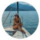 Frau mit Sonnenbrille und Cap sitzt im Bikini auf einem Segelboot, im Hintergrund ist ein See und Berge
