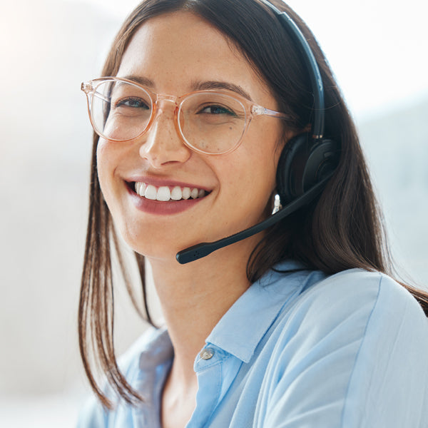Eine freundlich lächelnde Frau mit Brille trägt ein Headset und ein hellblaues Hemd.