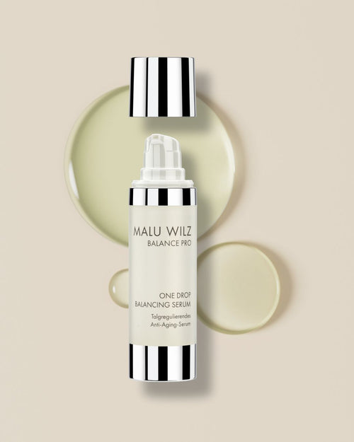 Malu Wilz Balance Pro One Drop Balancing Serum in einem Spender, umgeben von gelblichen Texturtropfen auf einem beigen Hintergrund.