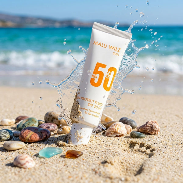 Eine Tube Malu Wilz Sun Protect Fluid mit LSF 50 steht im Sand vor einer Meereskulisse, wobei Wasser um die Tube spritzt und sie von Steinen und Muscheln umgeben ist