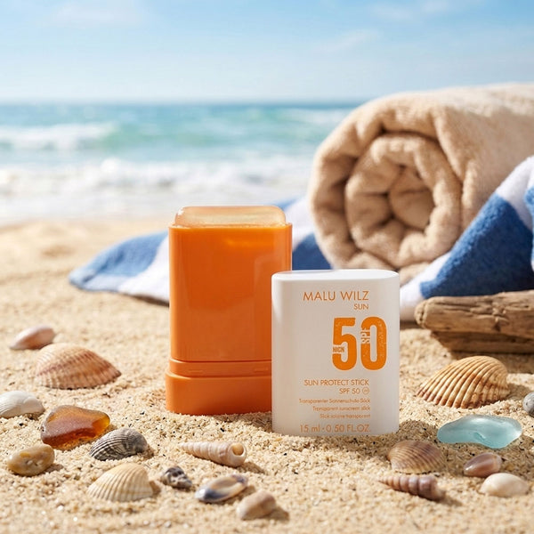 Ein transparenter Malu Wilz Sun Protect Stick mit LSF 50 steht am Strand im Sand, umgeben von Muscheln und einem Strandtuch vor der Meereskulisse