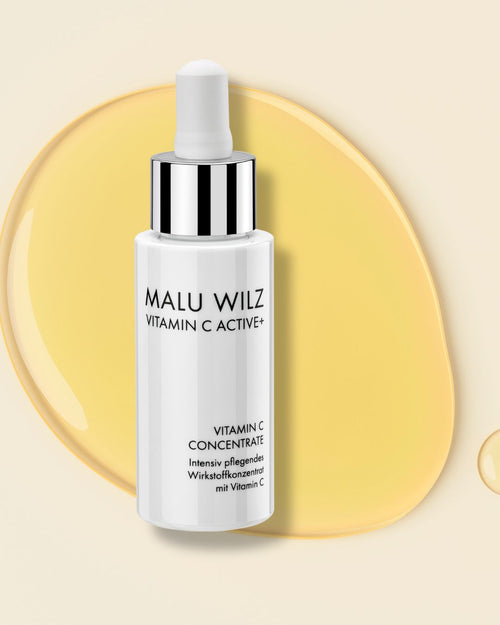 Malu Wilz Vitamin C Concentrate in einer weißen Pipettenflasche, platziert auf einem gelben Flüssigkeitstropfen.